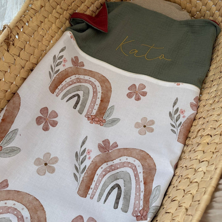 Babydeken met regenboog- en bloemenprint, groene achterkant en geborduurde naam in een mand.
