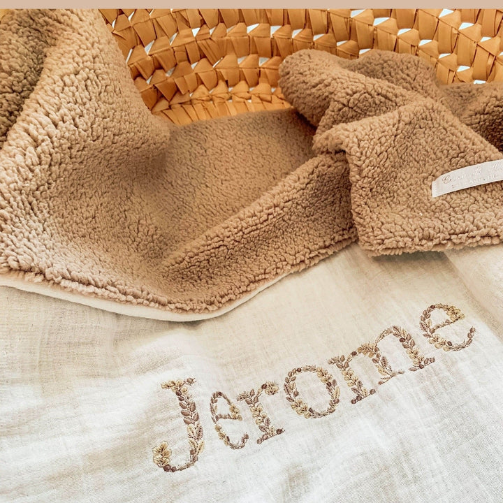 Beige babydeken van teddy en hydrofiel met geborduurde naam, opgevouwen in een mand.