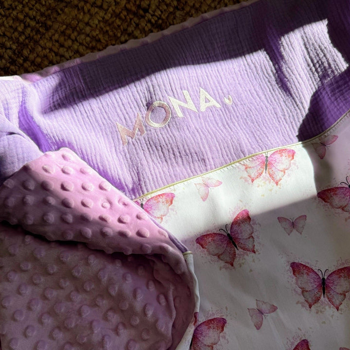 Babydeken met vlinderprint, lila achterkant en geborduurde naam, met roze noppenstof zichtbaar.