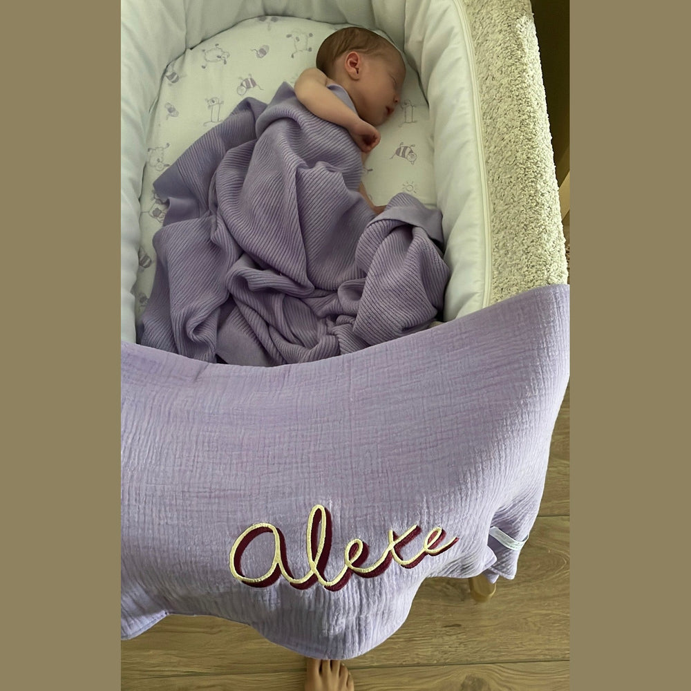 Baby ligt onder een lavendelkleurig hydrofiel babydeken met teddyachterkant.