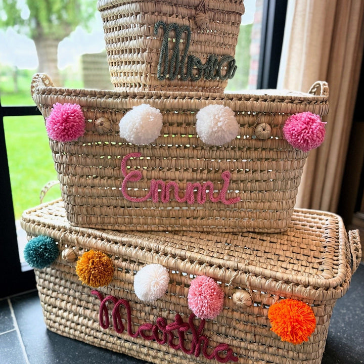 Twee palmbladmanden met namen en pompons in verschillende kleuren.