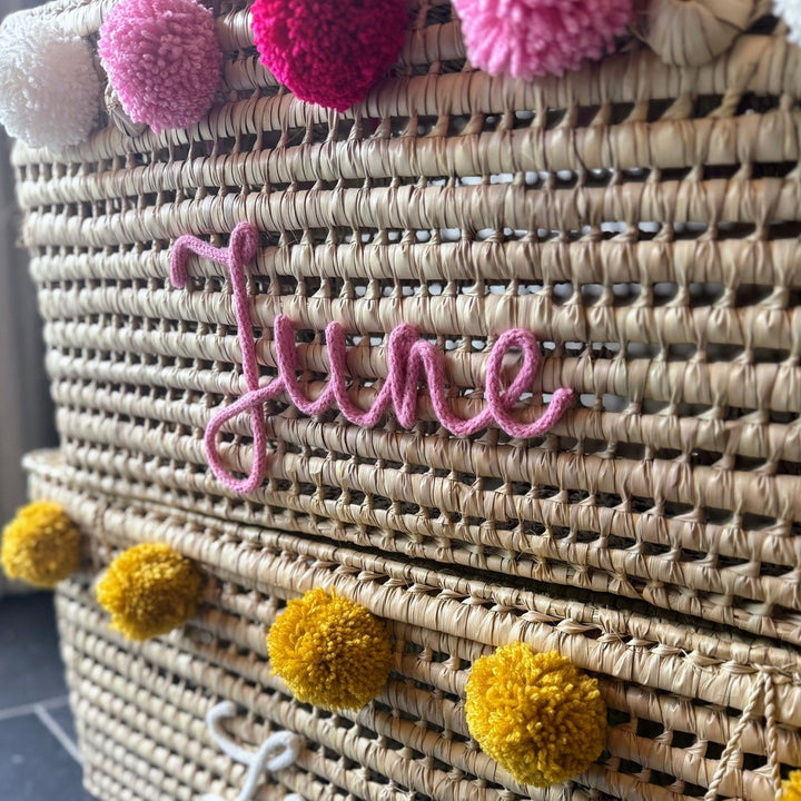 Palmbladmand met roze naam en pompons in zachte kleuren.