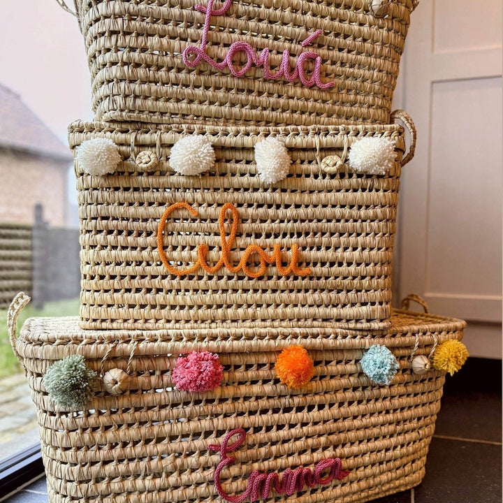 Zijaanzicht van palmbladmand met naam en rij kleurrijke pompons.